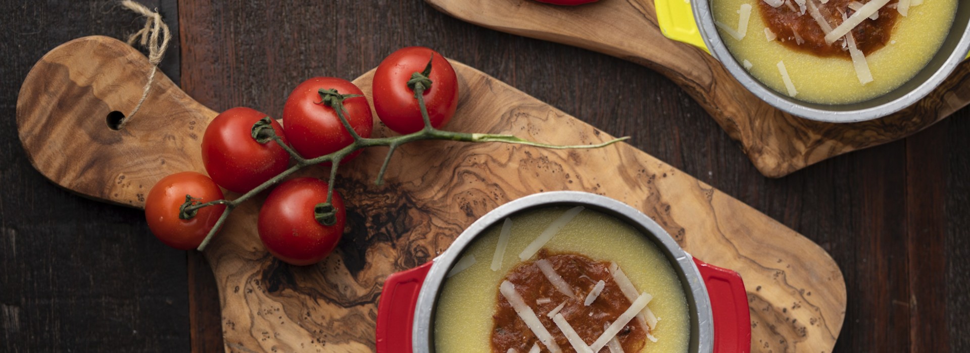 Polenta cremosa com molho e queijo servida em panelinhas, sobre tábua de madeira com tomates frescos