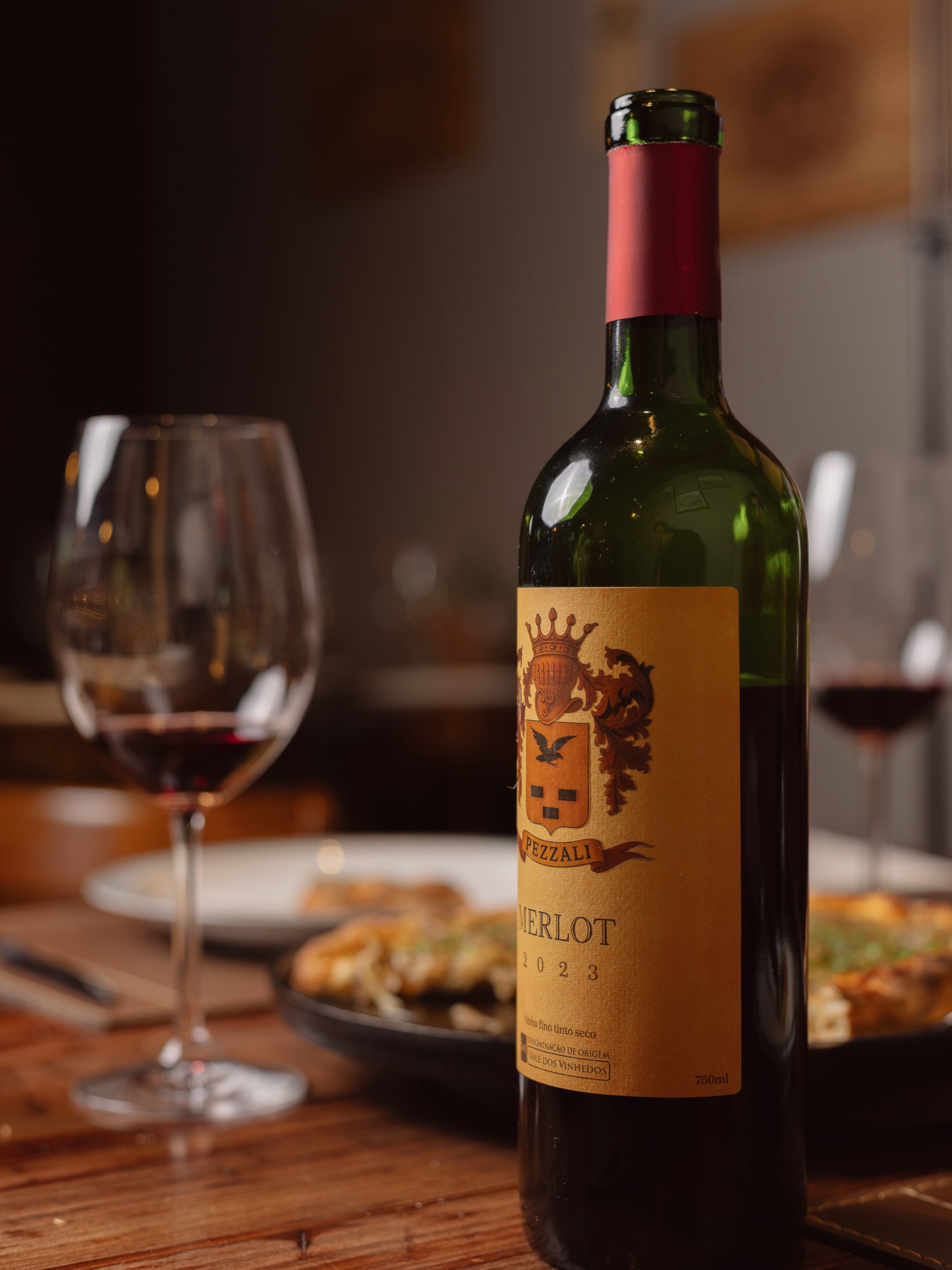 Garrafa de vinho tinto aberta sobre a mesa, com taças servidas ao fundo