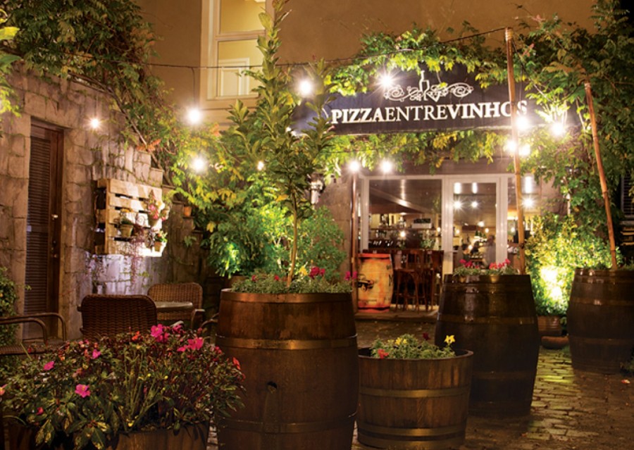 Pátio da Pizza Entre Vinhos à noite, com barris de vinho, plantas e luzes acesas