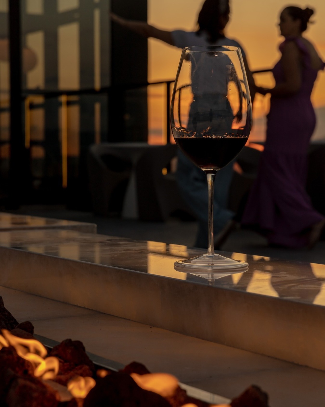 Taça de vinho tinto na borda da lareira do terraço, com duas pessoas em silhueta olhando o pôr do sol